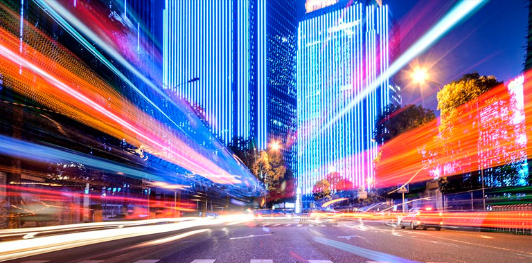 Cidade de noite com as luzes dos postes acesas e luzes de carros que passam