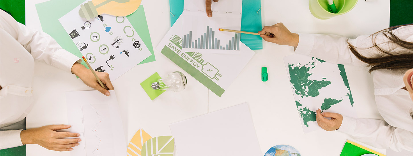 Três pessoas se reunem sob uma mesa com alguns papéis sob ela, um dos papéis tem gráficos duas pessoas apontam para ele, outro tem um mapa, folhas na cor bege, um ícone de lampada recortado e aaixo um papel com ícones diversos impressos na folha