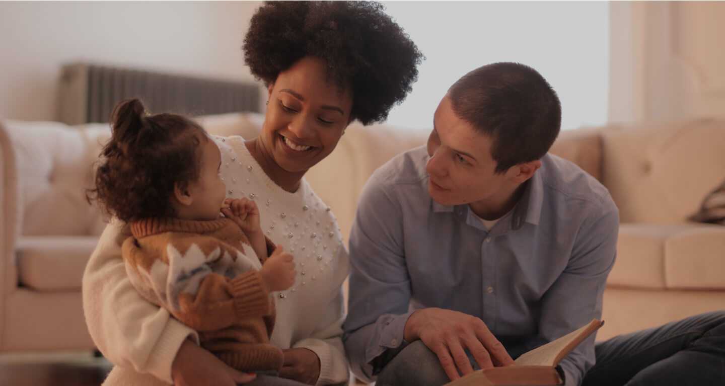 Família negra sentado no chão da sala de casa, a mãe sorri, contam uma história para sua filha de 2 anos