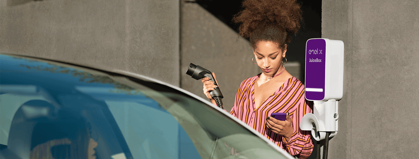 Mujer joven cargando un auto eléctrico con el equipo de Juice Box, mientras revisa su carga desde el celular. 