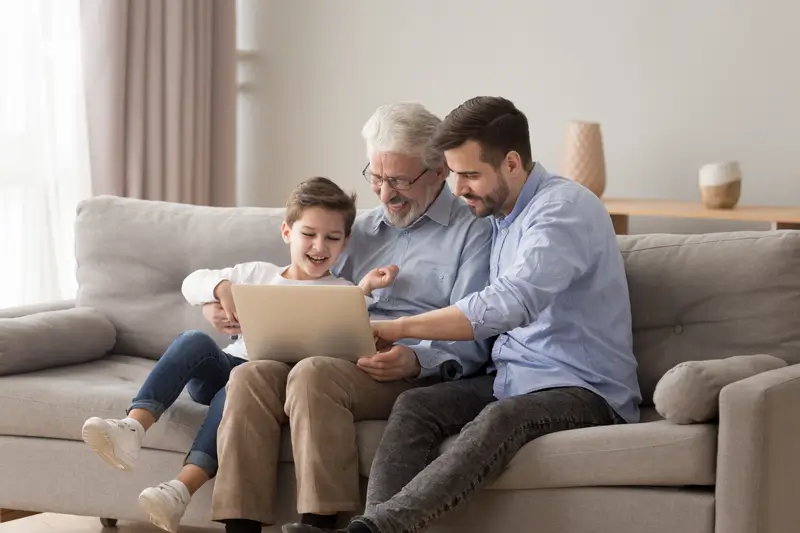 Padre, hijo y nieto sentados en un sofá, sonriendo frente a un computador, al revisar las características del plan de Asistencia Funeraria de Enel X.