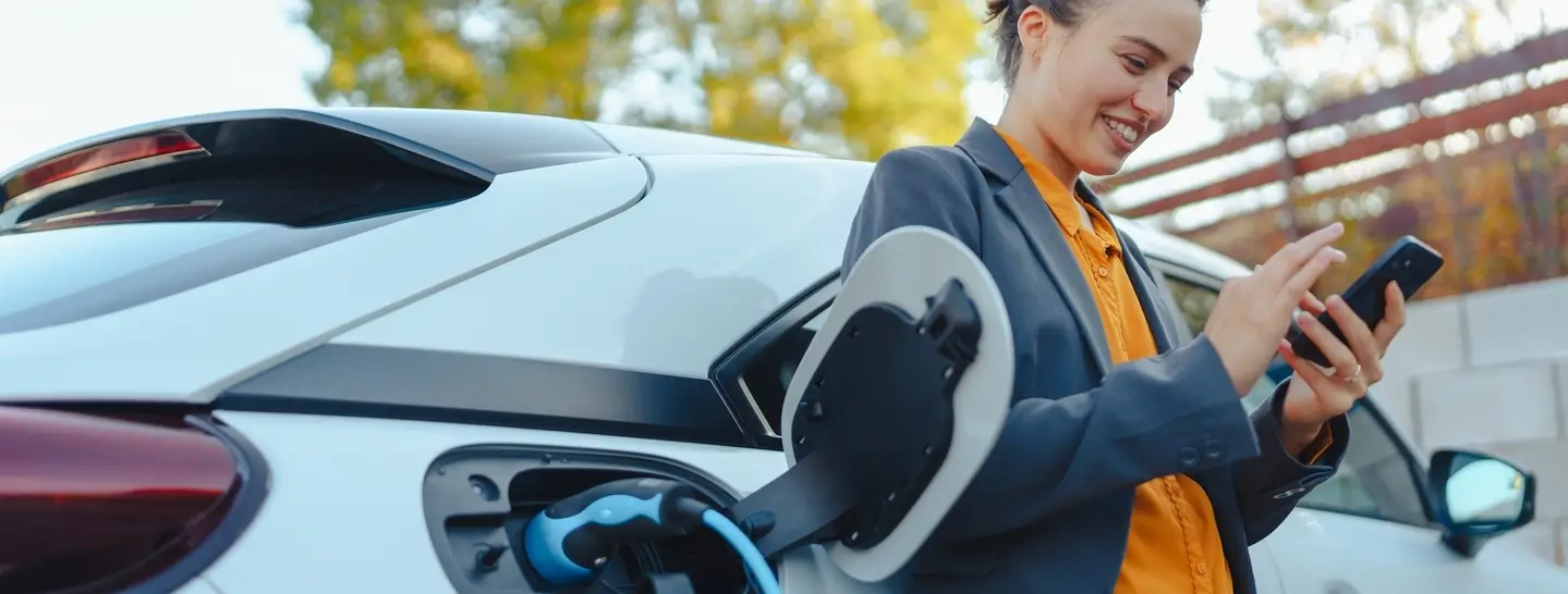 Mujer revisando en su celular el nivel de carga de su carro eléctrico, lo que destaca claramente la diferencia entre un vehículo eléctrico y uno de combustión.