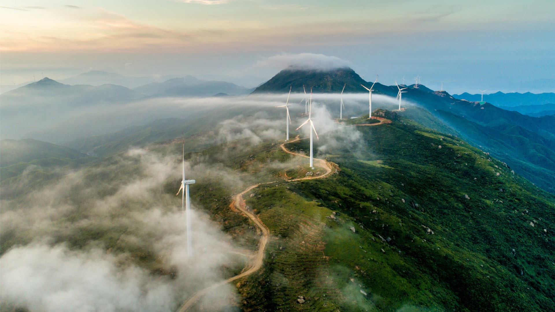 Renewable energy wind turbines atop of a hill with mist and cloud floating alongside.